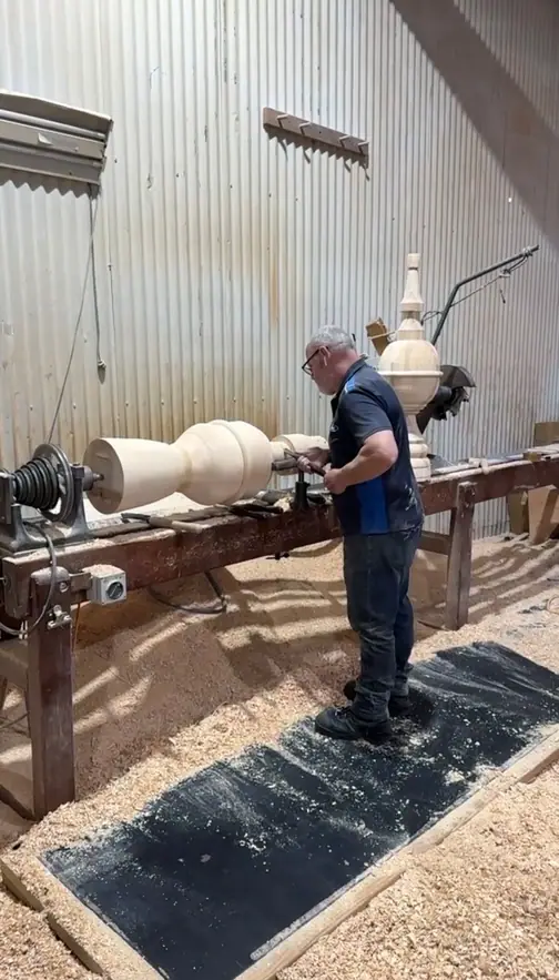 A turner working a length of pine on the long-bed lathe at Bayswater, wearing safety glasses, with a finished newel standing behind him.