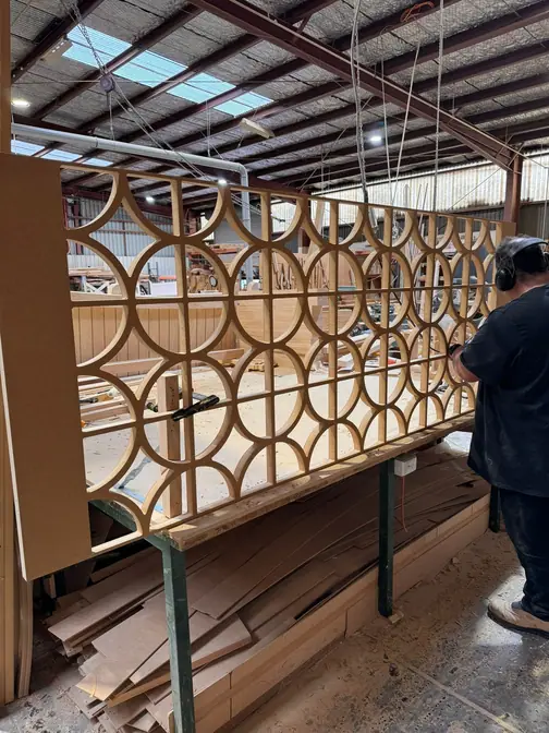 A worker in ear defenders standing beside a very large fretwork panel of interlocking circles, stood on edge in the Bayswater workshop.