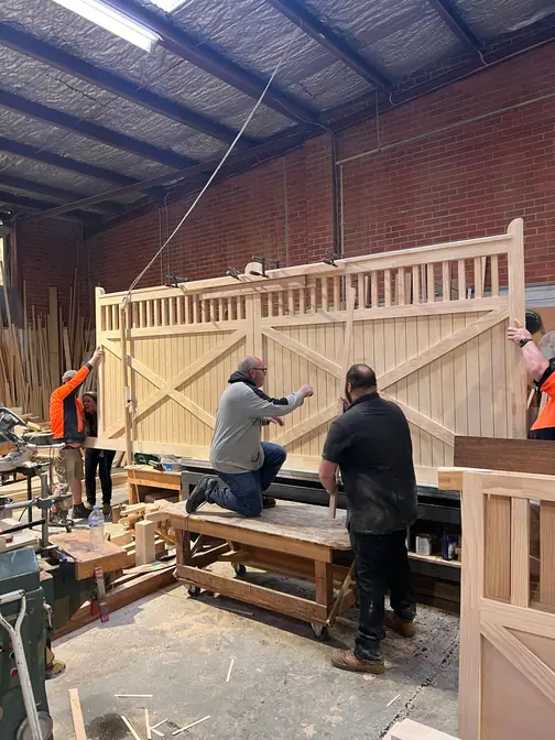 Three men assembling a large braced timber gate with a turned picket top on trestles inside the Bayswater workshop.