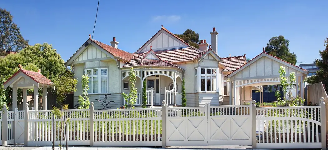 A cream Federation house photographed from the street: a roofed lych gate over the front path at the left, a picket fence with turned infill and X-braced driveway gates, a bay window, gabled roof, a verandah with timber fretwork and balustrade, and a gabled carport at the right.