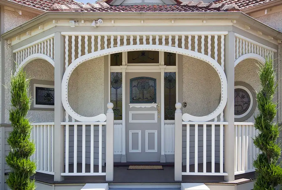 Close view of a rendered period house entry: a timber frieze runs under the verandah beam, a large shaped fretwork arch springs between the posts, and an assembled balustrade with ball-topped newels runs each side of a leadlight front door.
