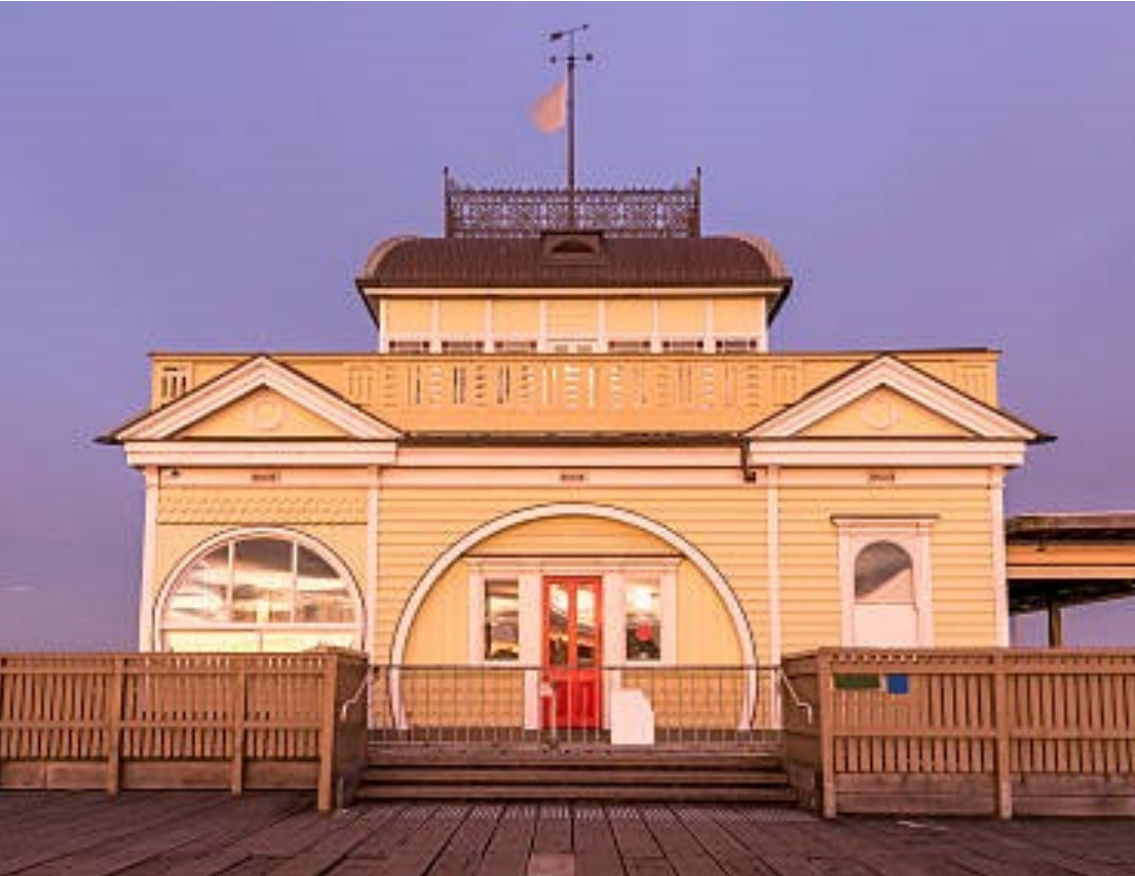 A weatherboard pavilion at dusk, its doorway set within a broad timber arch above the decking